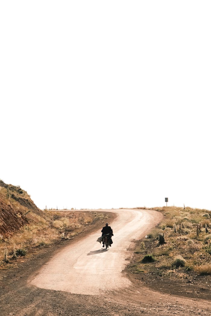 A solitary motorcyclist rides along a deserted dirt road in a barren landscape.