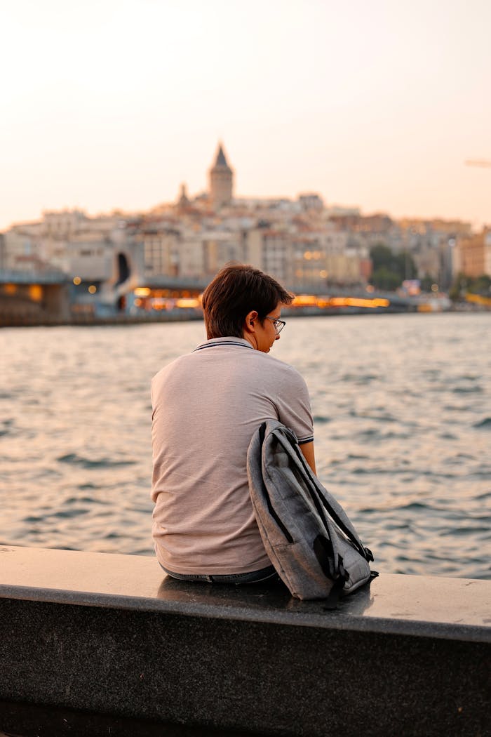 Man with a backpack sits at sunset by the water with Istanbul skyline view.