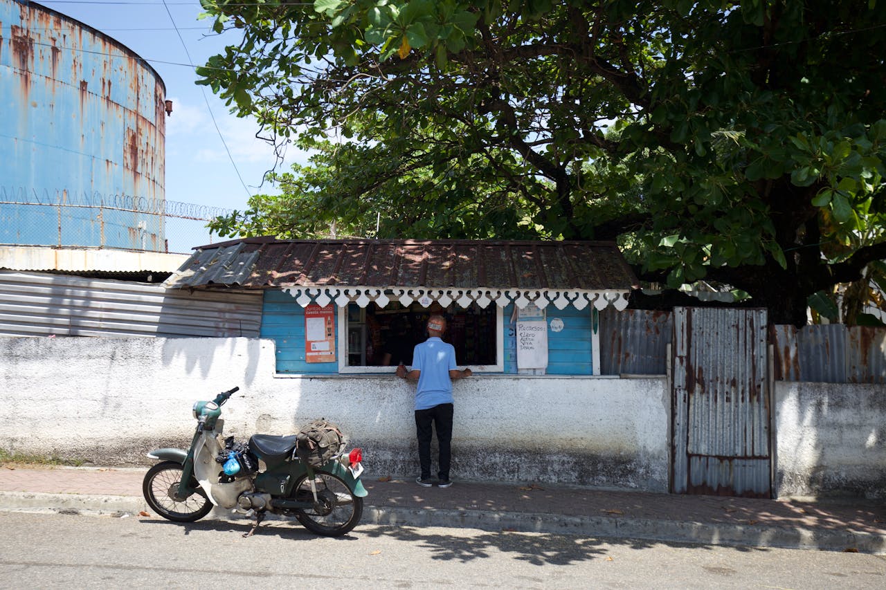 A street vendor stands by a small shop and parked scooter under a tree in the Dominican Republic.